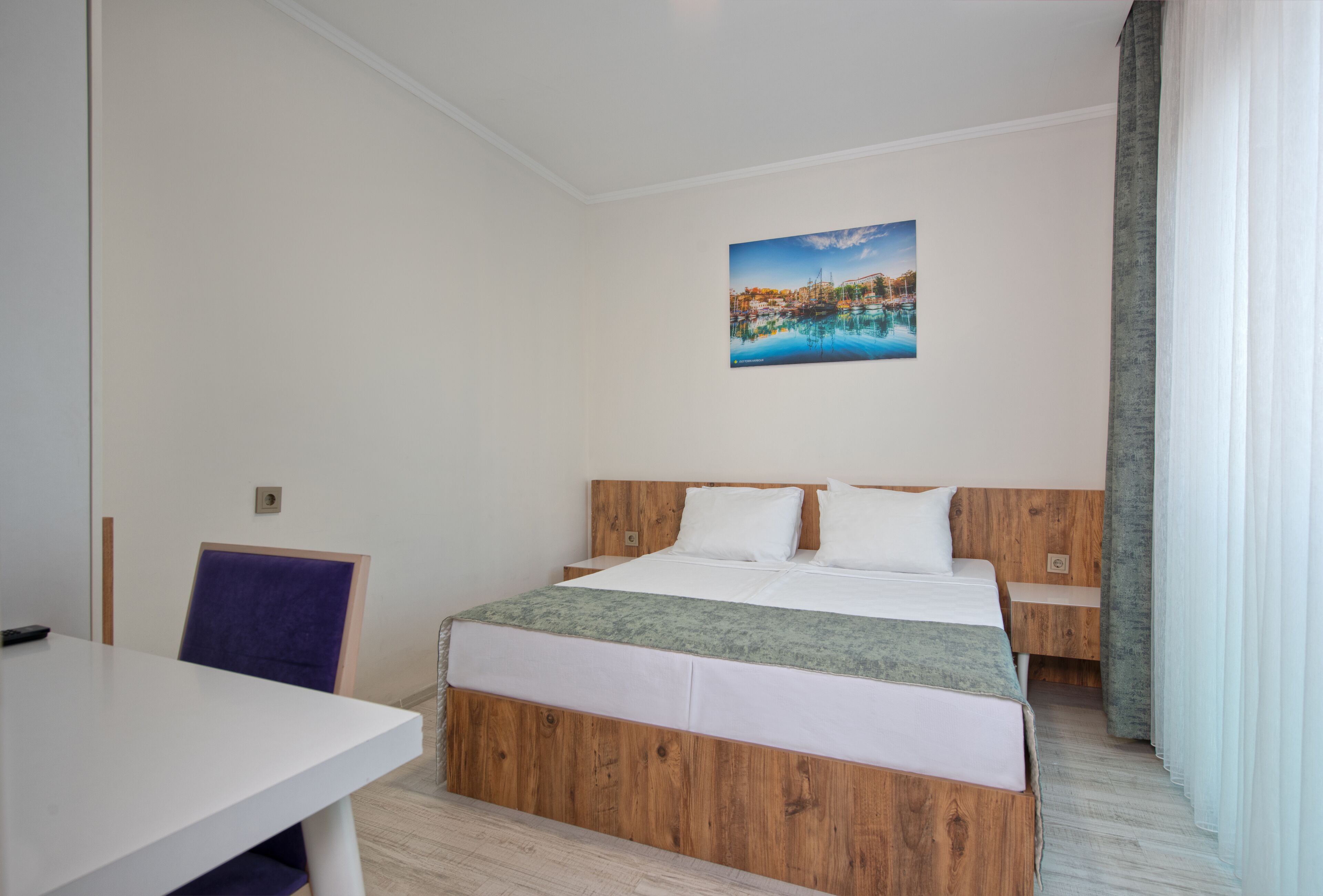 A hotel room with a wooden bed, white bedding, a small table, a chair, and a painting of a coastal scene on the wall.