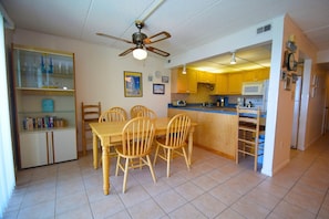 A dining area with a wooden table and chairs, a ceiling fan, and a kitchen with yellow cabinets and a microwave. - Thunder Island 80b 2 Bedroom Condo by RedAwning (Ocean City)