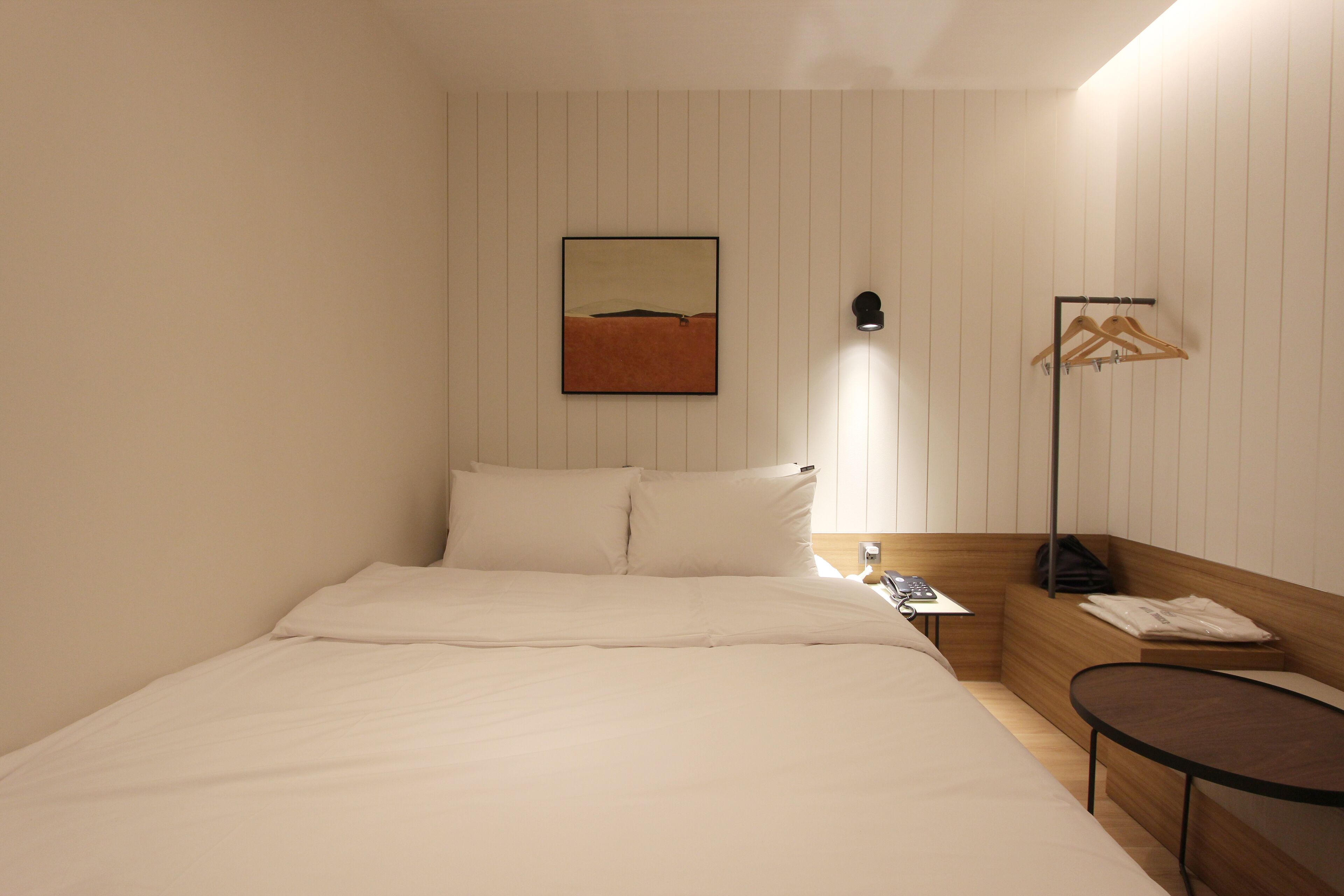 Une chambre d’hôtel avec un lit, une table de chevet avec une lampe, un porte-manteau et un tableau accroché au mur.