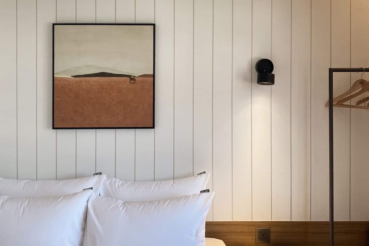 Una habitación de hotel con cama, almohadas, un cuadro enmarcado en la pared, un perchero y una lámpara.
