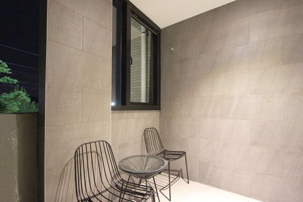 Un espace extérieur moderne, aménagé avec deux chaises en métal et une table en verre, le tout placé devant un mur sobre, avec une fenêtre au-dessus.