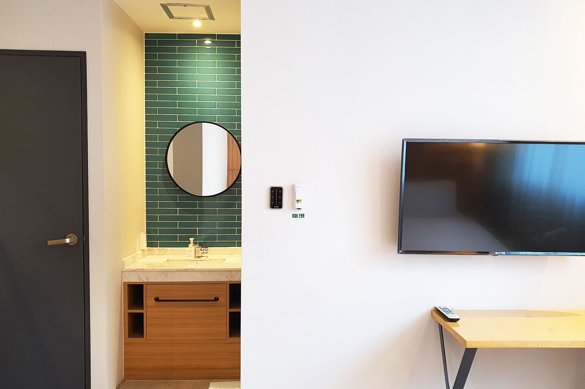 A modern bathroom with a green-tiled wall, a round mirror, a sink with a faucet, a toilet, and a flat-screen TV mounted on the wall.