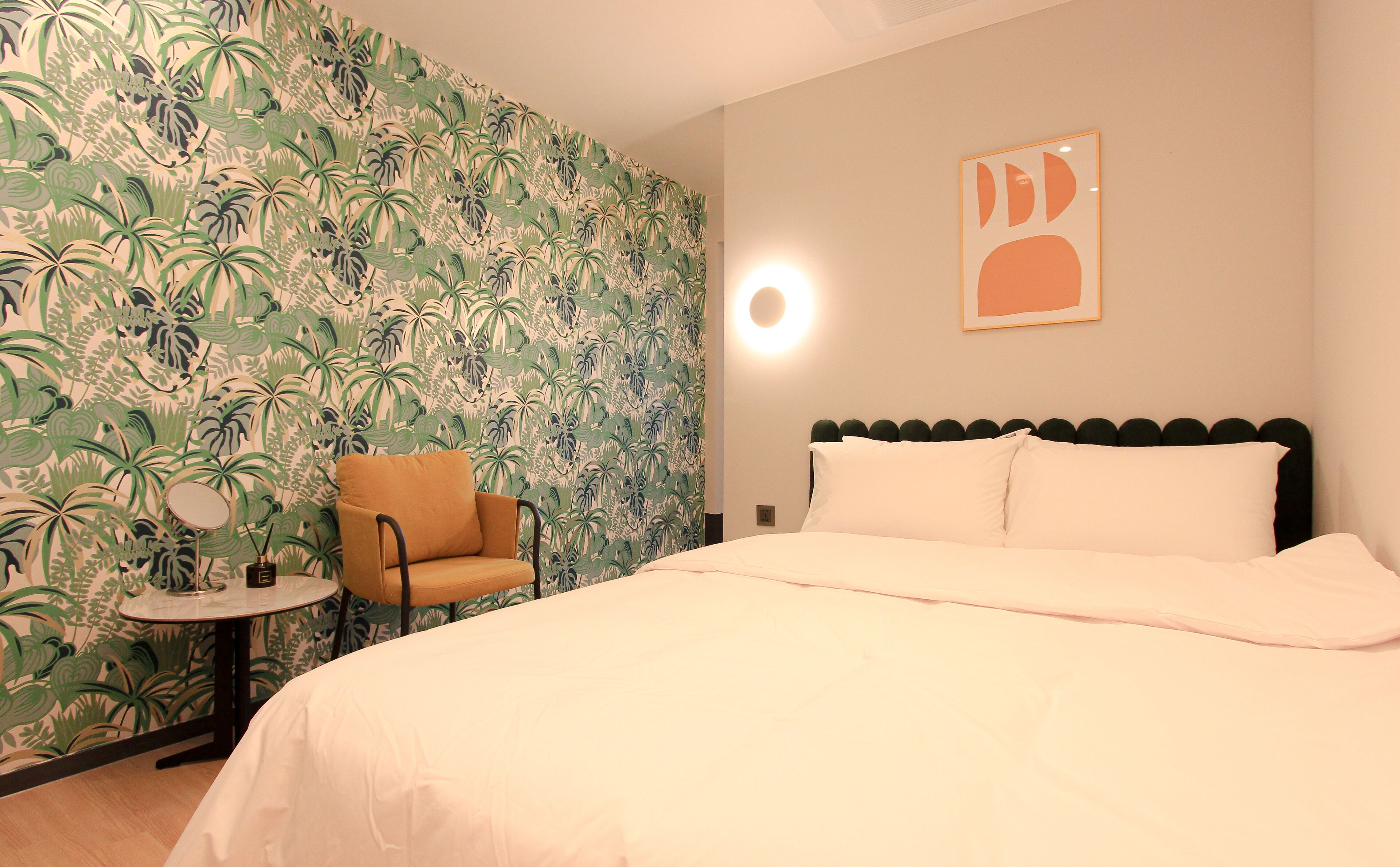 A hotel room with a bed, a chair, a small table, and a wall with a tropical leaf pattern.