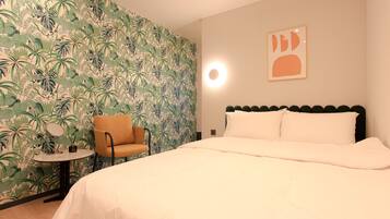 A hotel room with a bed, a chair, a small table, and a wall with a tropical leaf pattern.