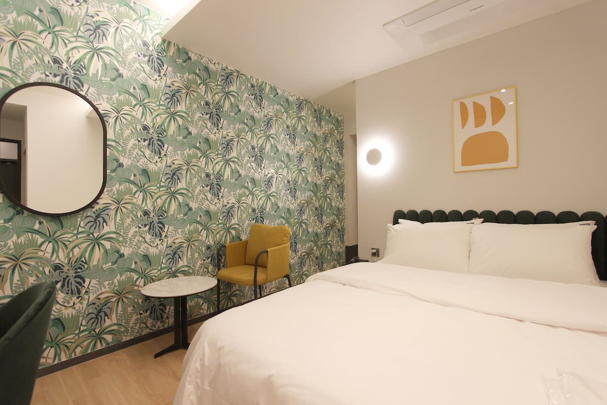 Habitación de hotel con cama, un espejo redondo, una mesita pequeña y redonda, una silla amarilla y un papel tapiz tropical.