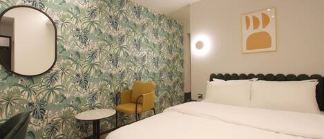 A hotel room with a bed, a round mirror, a small round table, a yellow chair, and a tropical wallpaper.