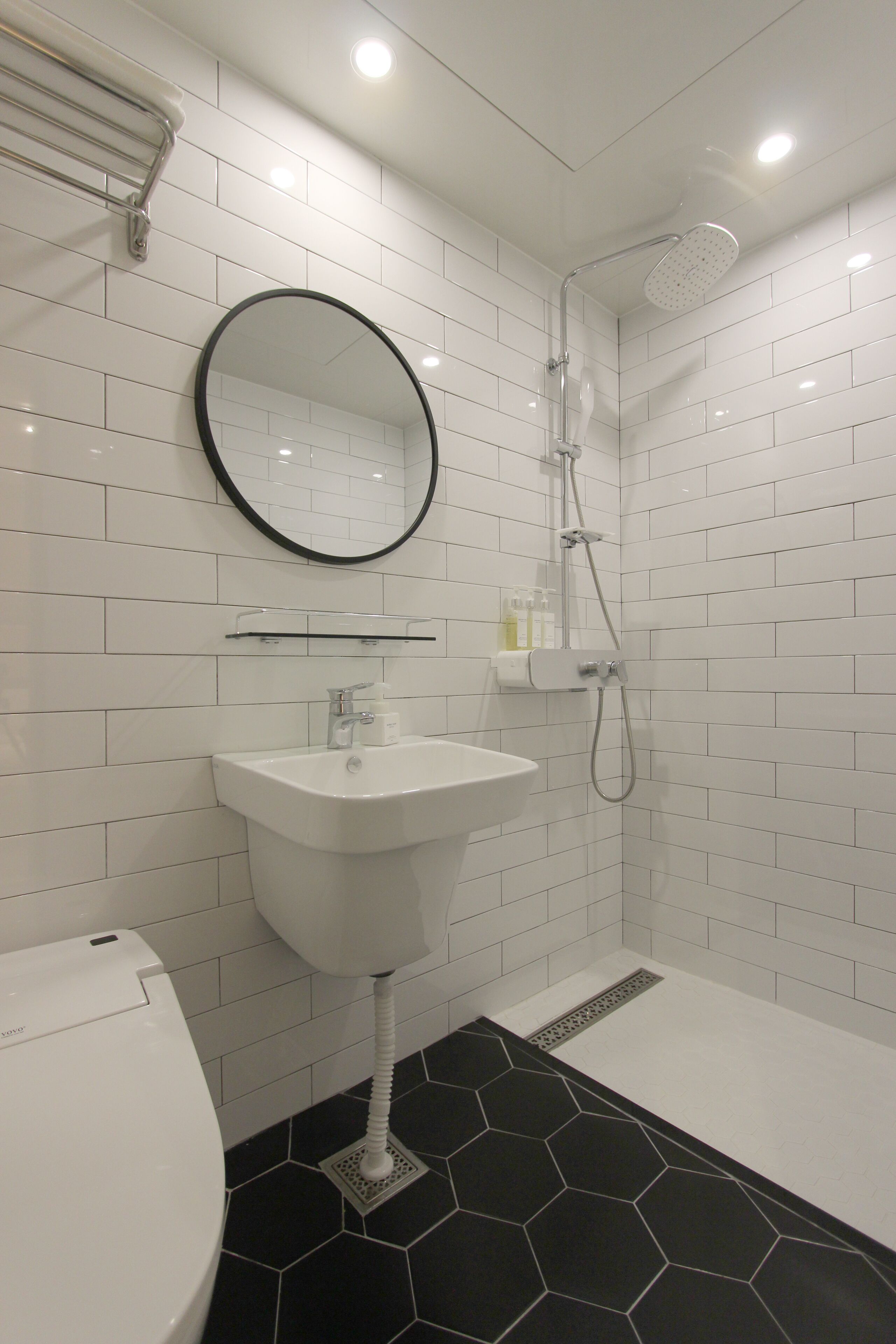 A bathroom with white tiled walls, a black hexagonal floor, a white sink, a round mirror, and a shower with a handheld showerhead.