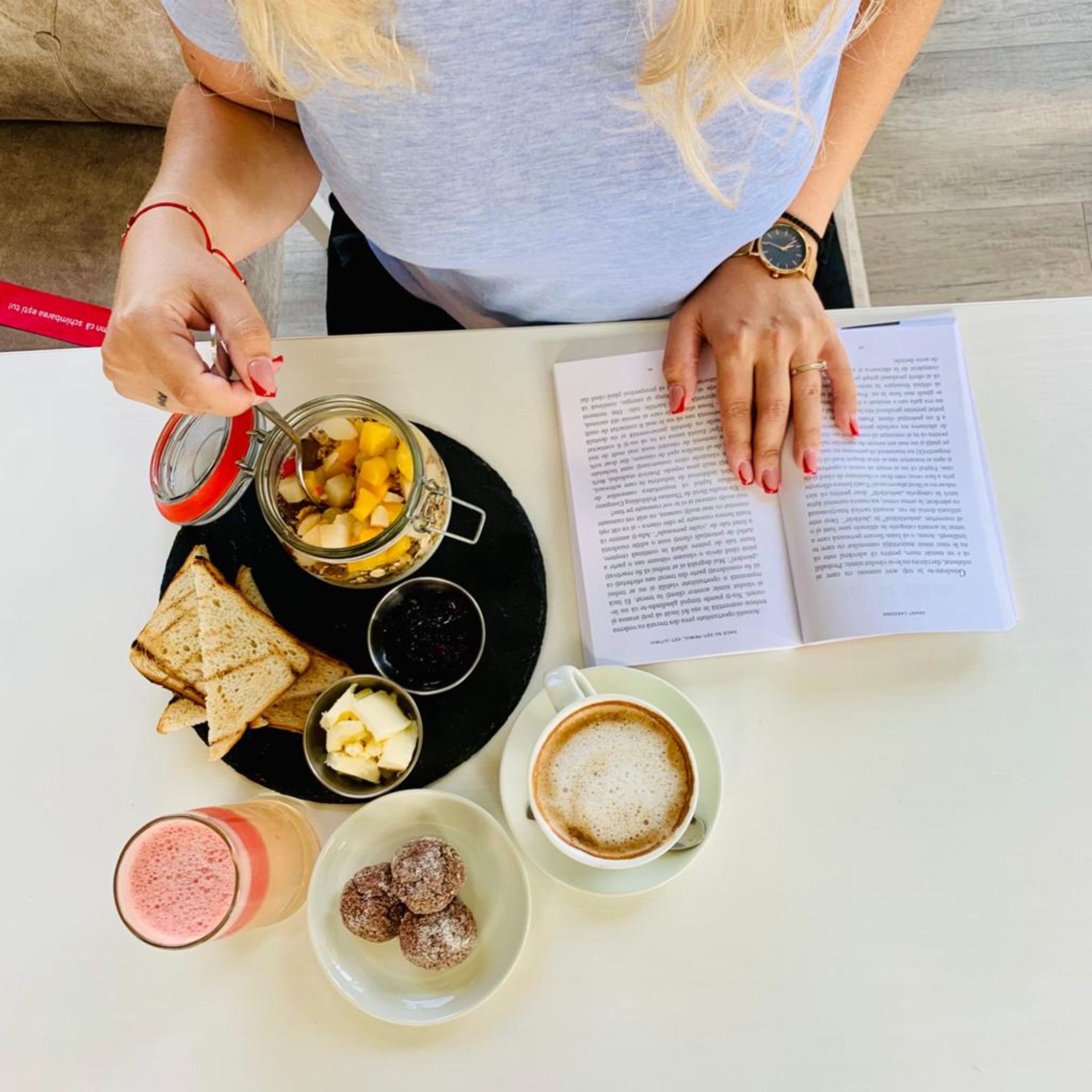 A person sitting at a table with a book, a glass of coffee, a jar of fruit, a plate with toast and butter, and a glass of smoothie.