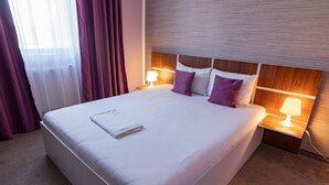 Una cama bien tendida con sábanas blancas y almohadas moradas, un cabecero de madera y lámparas de noche.
