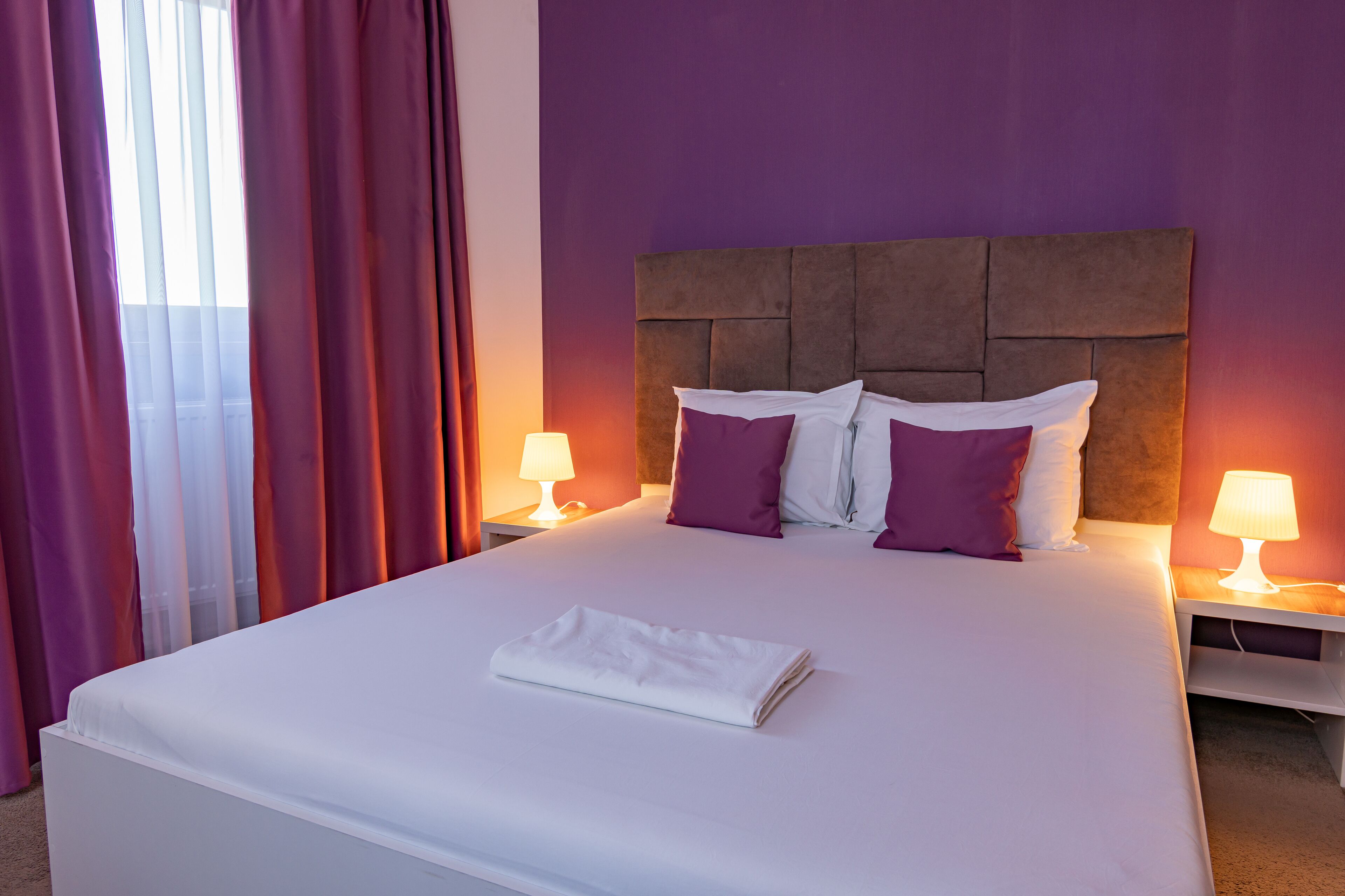 A neatly made bed with white linens and purple pillows, a brown headboard, and two bedside lamps on a white nightstand.