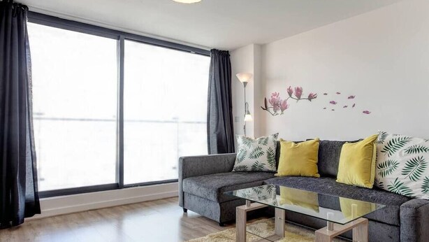 A modern living room with a grey sofa, yellow cushions, a glass coffee table, and a large window with curtains. - Clean and modern Riverside Flat w/ Private Balcony (Norwich)