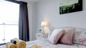 A hotel room with a bed, pillows, a nightstand, a lamp, and a window with curtains. - Clean and modern Riverside Flat w/ Private Balcony (Norwich)