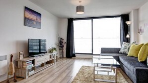 A modern living room with a flat-screen TV, a glass coffee table, a grey sofa with yellow cushions, a large window with black curtains, and a painting on the wall. - Clean and modern Riverside Flat w/ Private Balcony (Norwich)