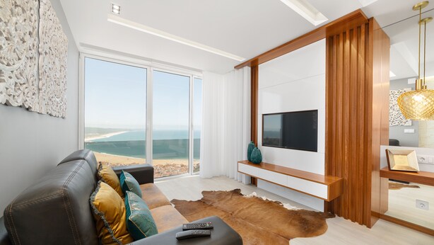 A modern living room with a leather sofa, a large flat-screen TV, and a view of the ocean through floor-to-ceiling windows. - Stunning View Apartment - Adults Only (Nazaré)