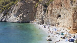 A beach with clear turquoise water, rocky cliffs, and several tents set up along the shore. - Agistri Apartments (Agistri)