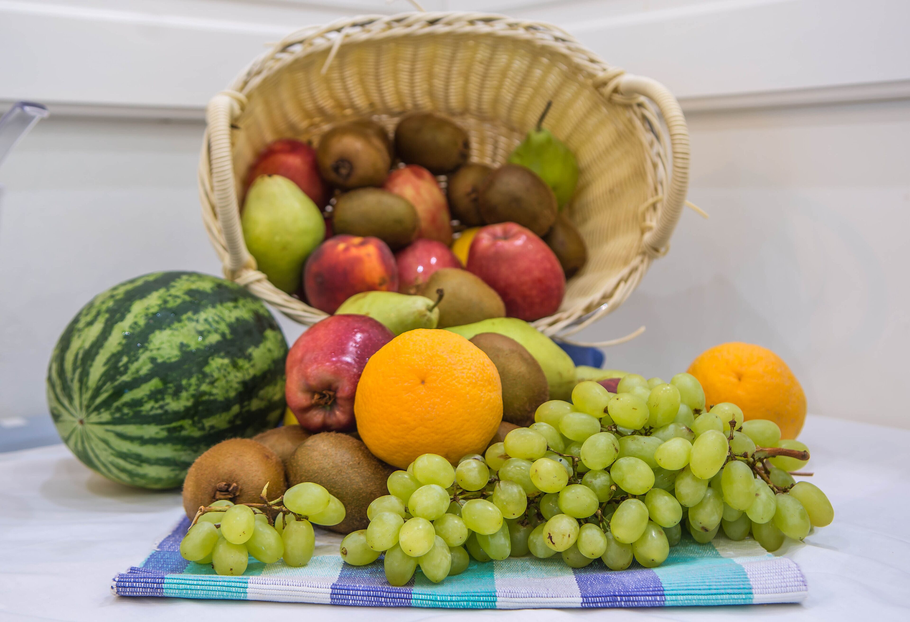 Uma cesta com frutas variadas, incluindo maçãs, peras, kiwis e laranjas, colocada sobre uma mesa com toalha xadrez.