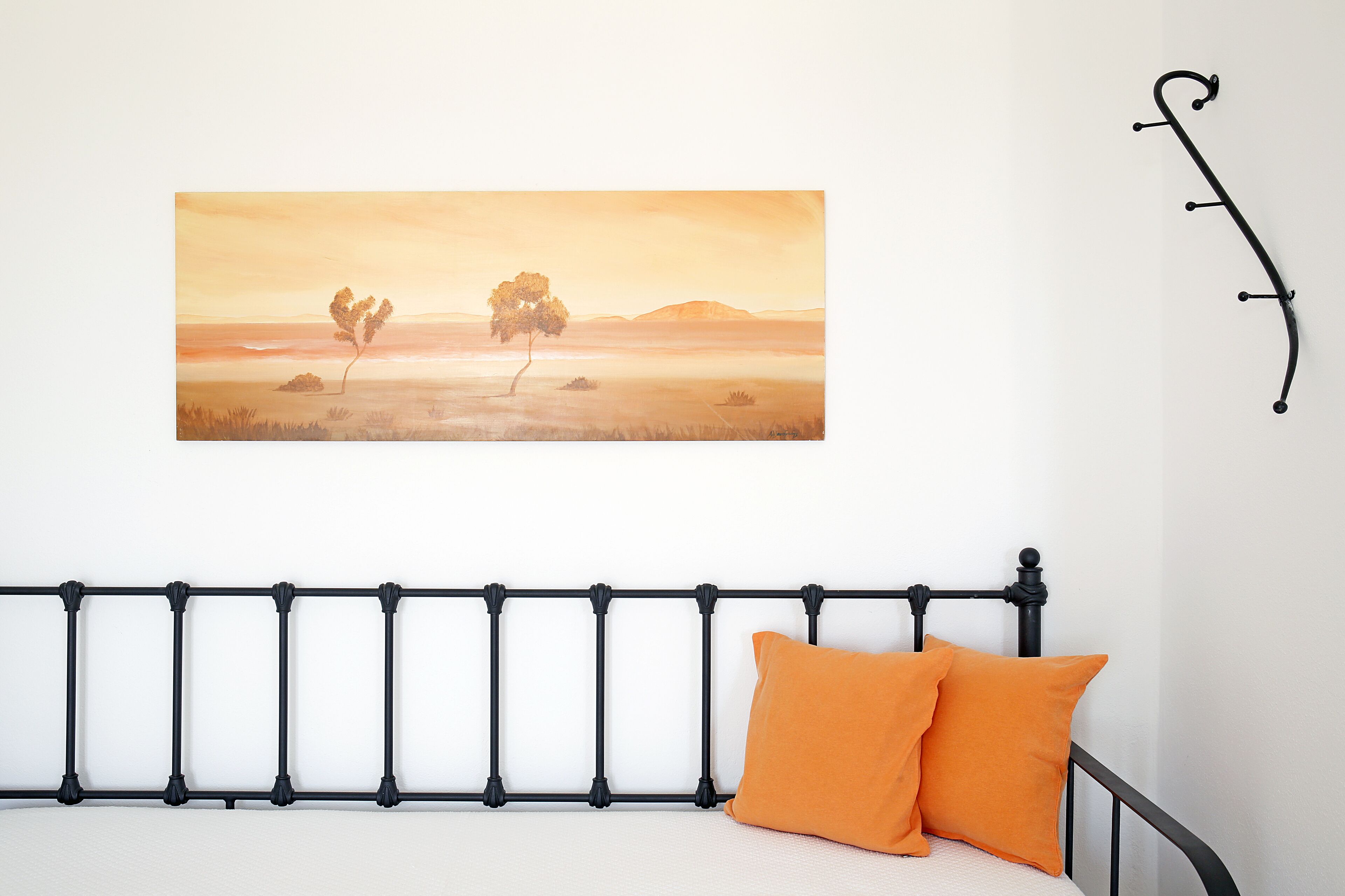 Una cama con cabecera de metal negro, dos almohadas naranjas y un cuadro de paisaje en la pared.