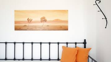 Una cama con cabecera de metal negro, dos almohadas naranjas y un cuadro de paisaje en la pared.