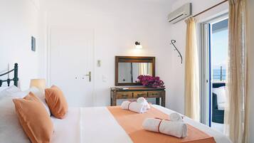 A hotel room with a bed, pillows, a bedside table with a mirror, a lamp, and a view of the sea through a window.