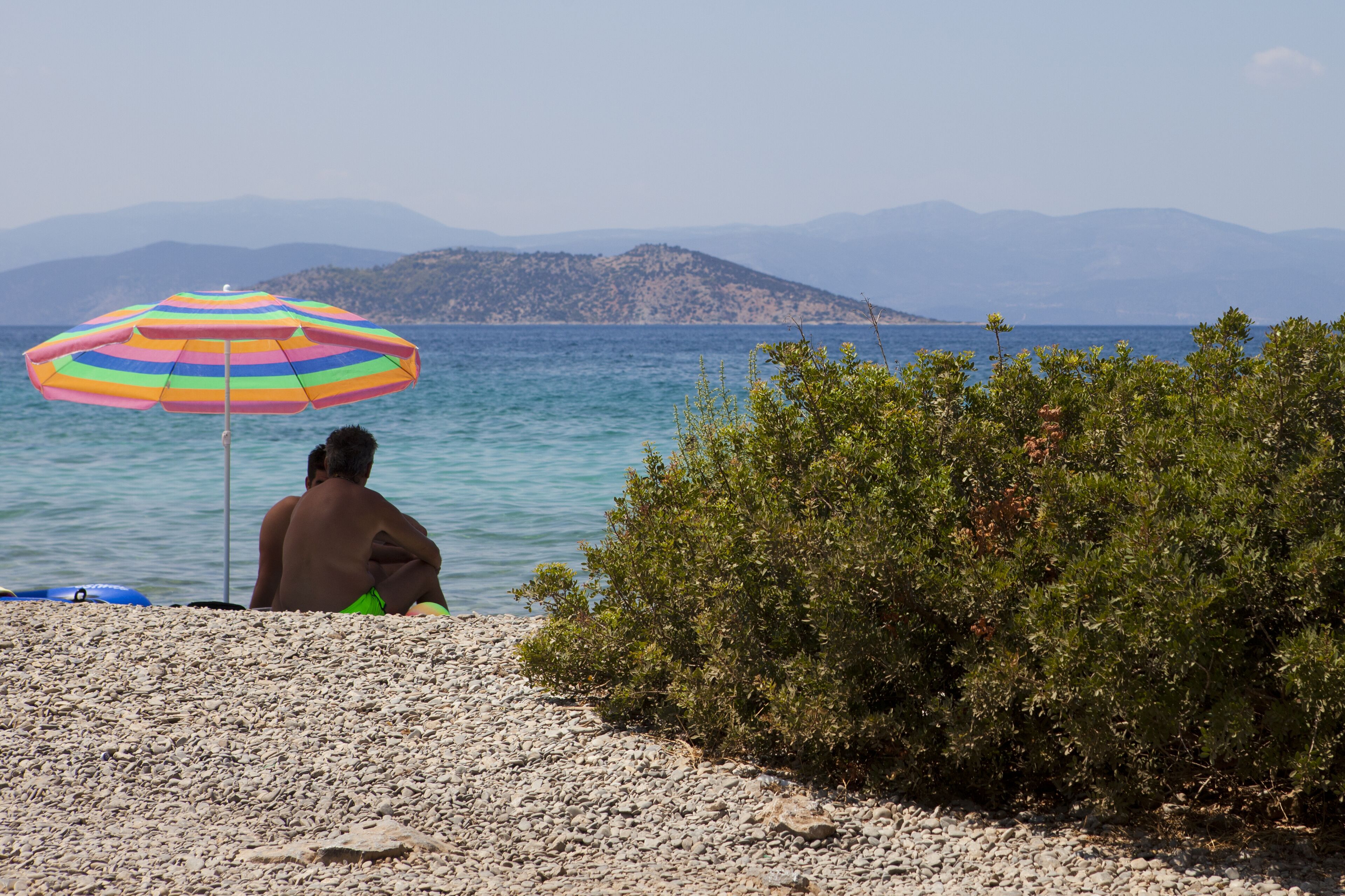 A beach with a person under a colorful umbrella, rocky terrain, and distant mountains.
