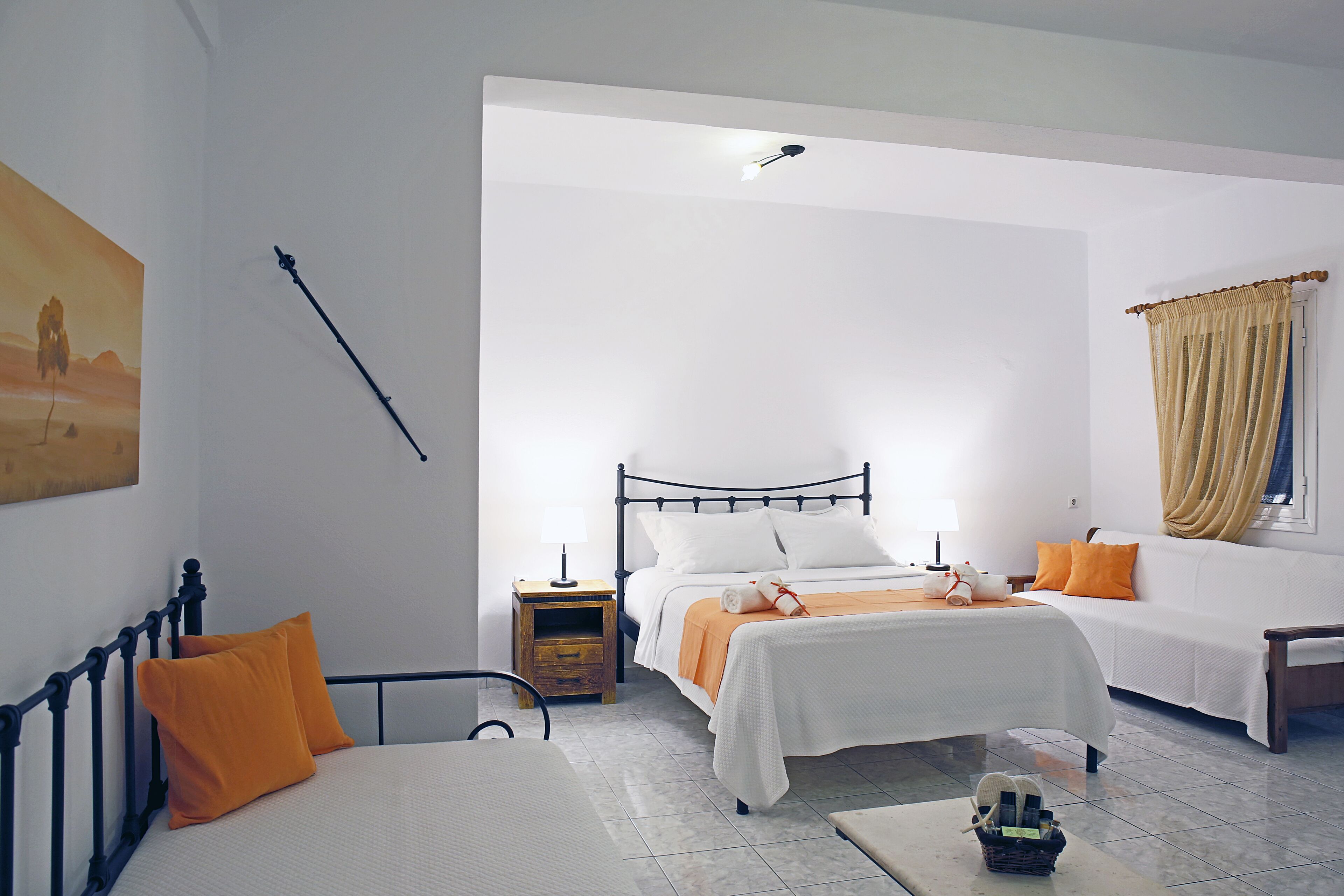 A hotel room with a large bed, a smaller bed, a desk, and a chair. There is a nightstand with a lamp, a painting on the wall, and a window with curtains.