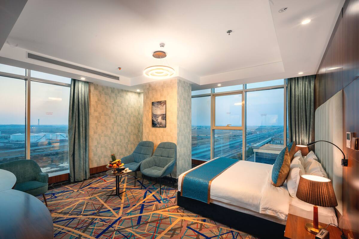 A hotel room with a large window, a bed, two chairs, a small table with fruit, and a lamp.