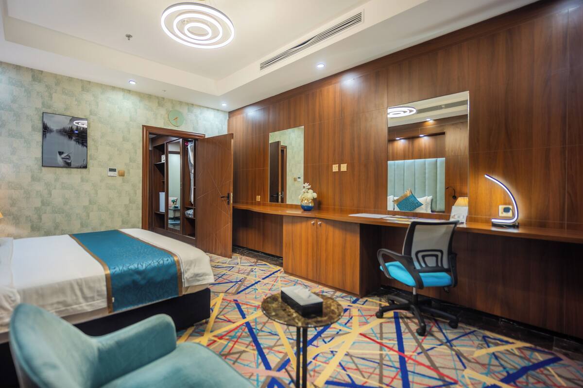 A hotel room with a bed, a desk, a chair, a mirror, and a patterned carpet.