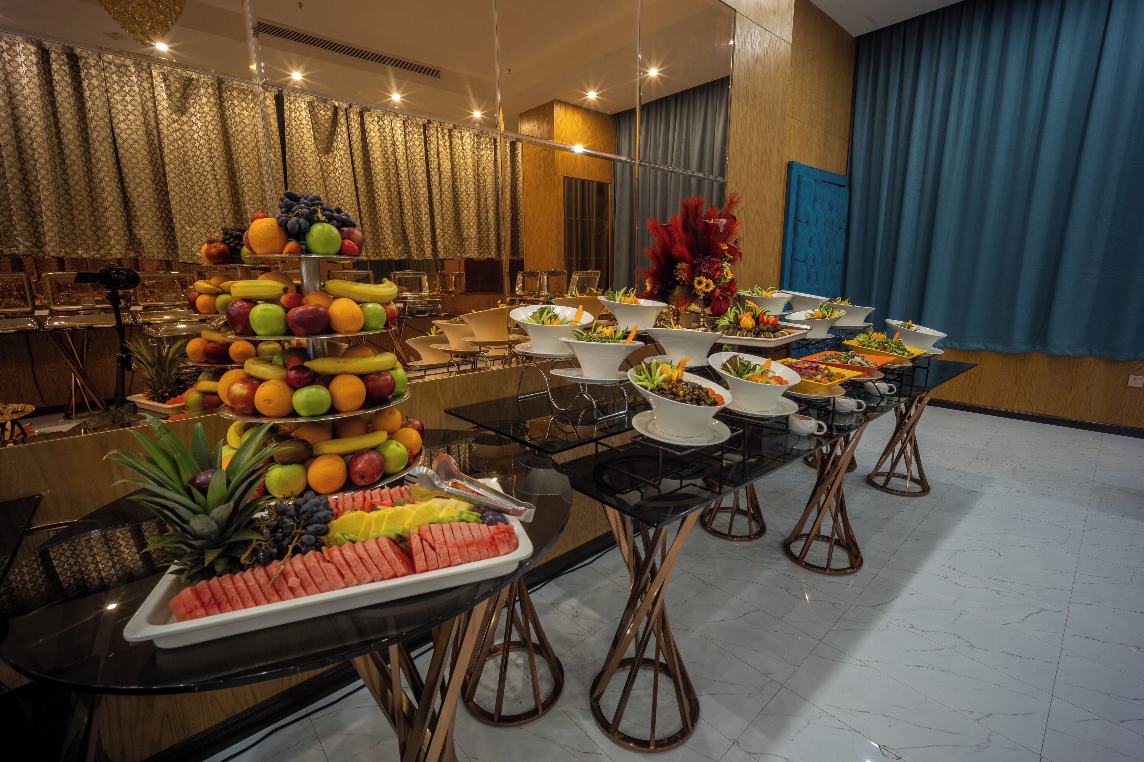 A buffet setup with various fruits and vegetables, a chandelier, and a blue curtain in a dining area.