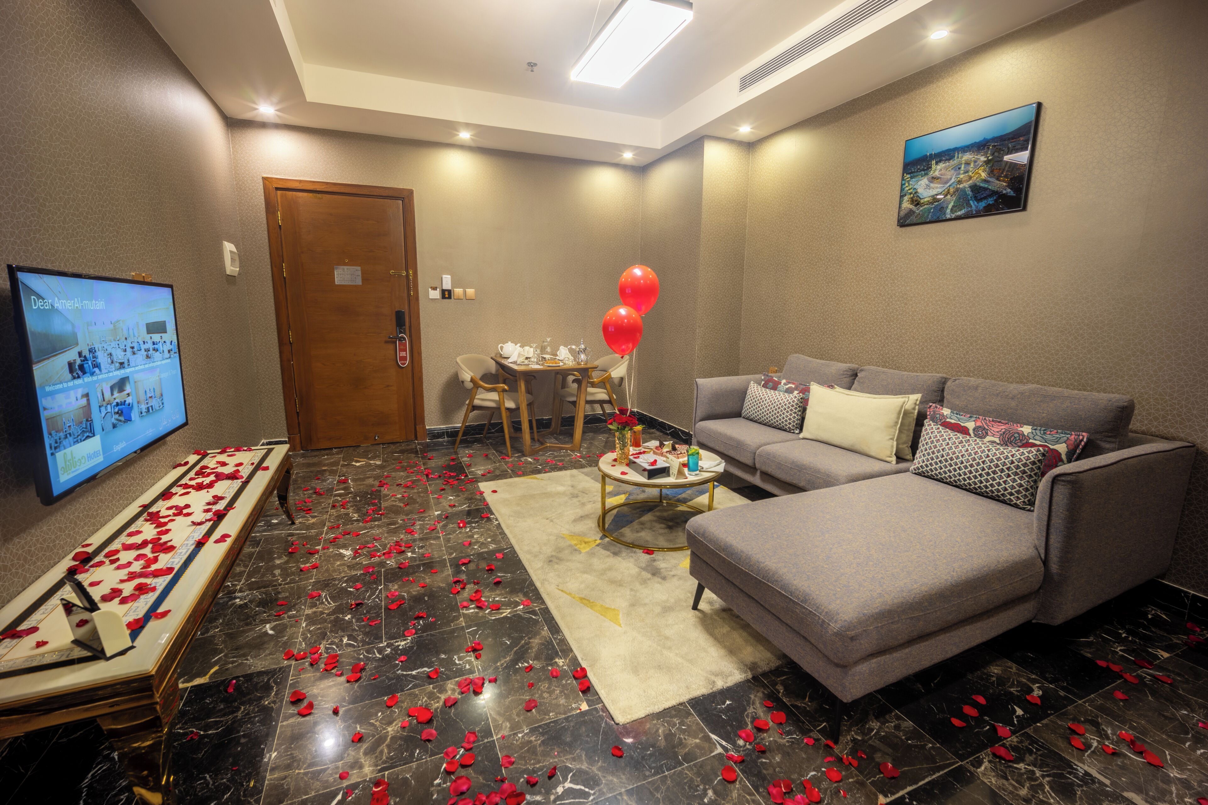 A hotel room with a sofa, a television, a dining table with chairs, and a carpet with rose petals.