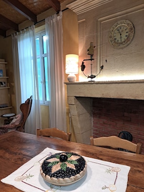 Et spiseområde med et træbord, stole, en dekorativ tærte på en underlag, en pejs, et ur og en lampe.