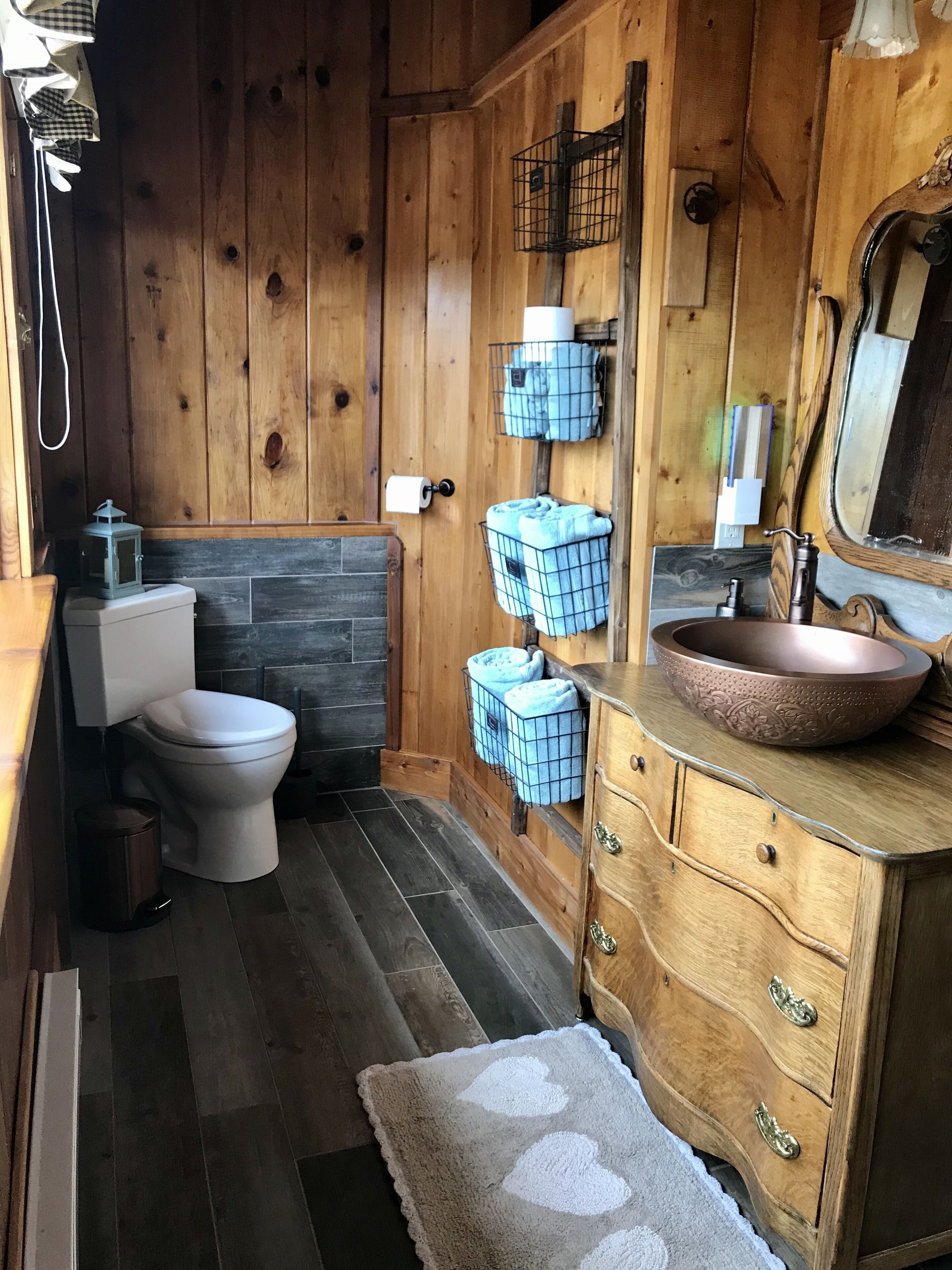 A wooden bathroom with a white toilet, a wooden vanity with a copper sink, and a mirror. There are two towel racks with folded towels, a trash can, and a small rug on the floor.