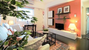Un salon avec un canapé, une table basse et un téléviseur placés contre un mur rouge. Une plante est accrochée au mur et une lampe se trouve sur une table d’appoint.