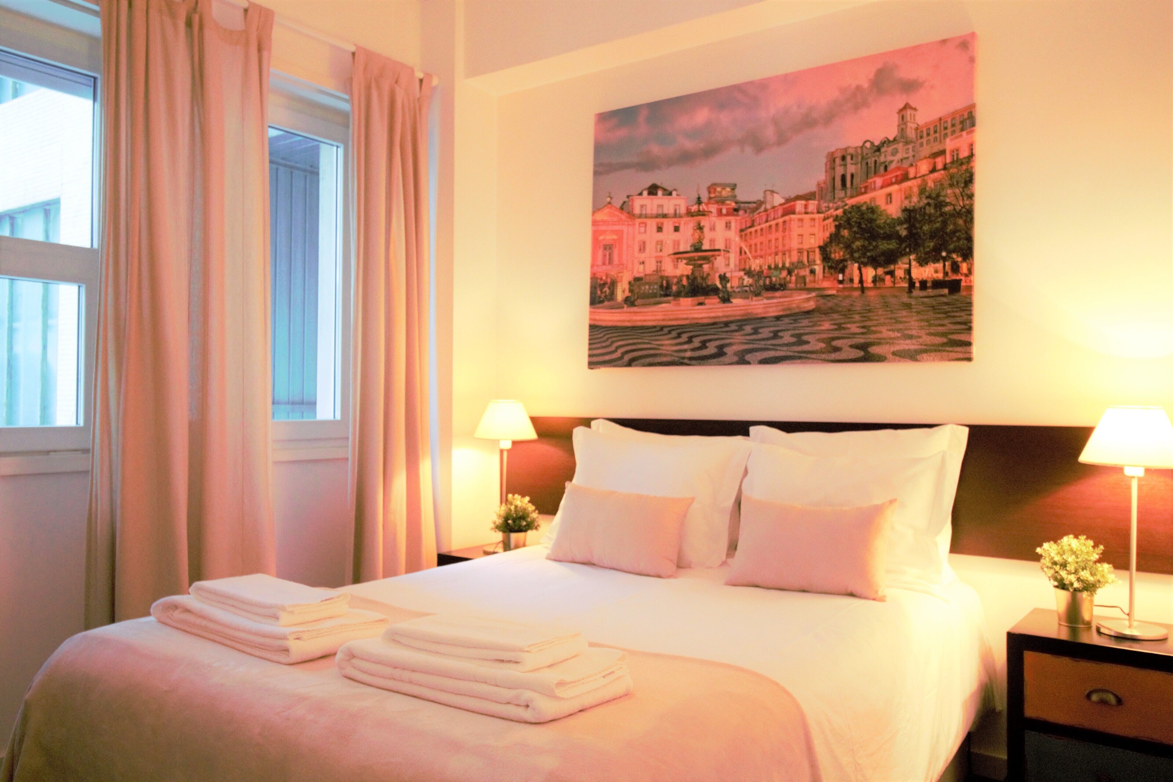 A neatly made bed with folded towels, a painting of a cityscape on the wall, and two bedside lamps.
