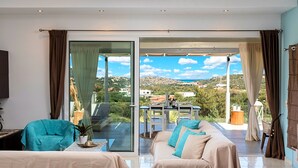 Un salon moderne avec un canapé et des fauteuils, une table à manger et une vue sur une colline et la mer, visible à travers de grandes portes vitrées.