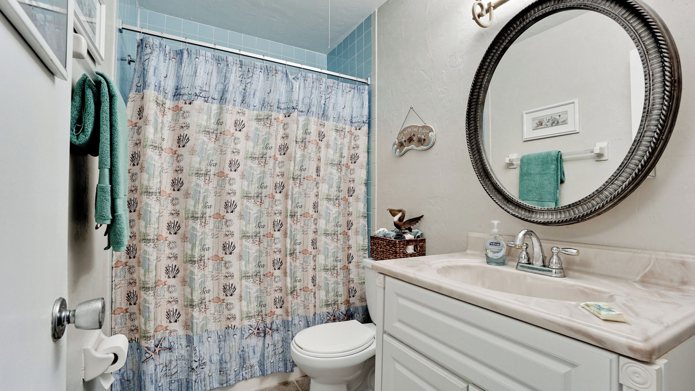 Une salle de bain avec un rideau de douche orné de motifs marins et de poissons, des toilettes blanches, un lavabo avec un distributeur de savon et un miroir doté d’un cadre décoratif.