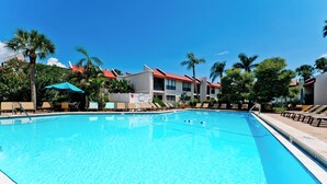 A clear swimming pool with lounge chairs and umbrellas, surrounded by palm trees and buildings with red roofs. - Runaway Bay 213 - 1845191 (Bradenton Beach)