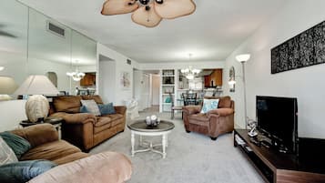 A living room with a sofa, armchairs, a coffee table, and a TV. There is a dining area in the background with a table and chairs.