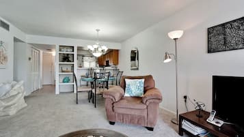 A living room with a beige sofa, a dining area with a table and chairs, a kitchen in the background, and a large floor lamp.