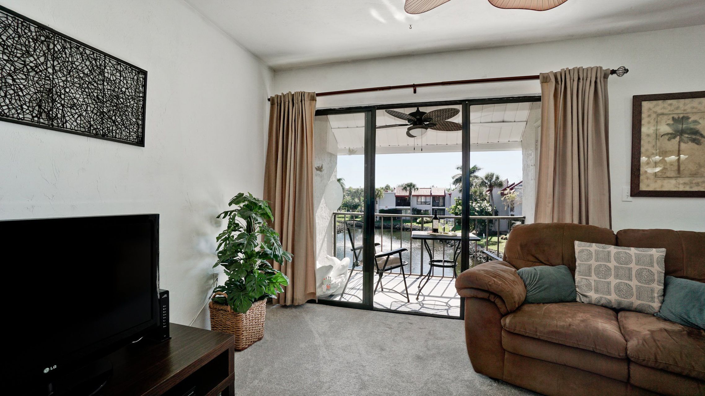 Uma sala de estar com televisão de tela plana, um sofá marrom com almofadas estampadas, uma planta em vaso e uma porta de vidro deslizante que dá para uma varanda com móveis de exterior.