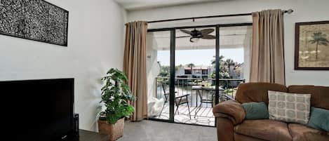 Una sala de estar con televisor de pantalla plana, un sofá marrón con cojines estampados, una planta en maceta y un ventanal corredero que da a un balcón con mobiliario exterior.