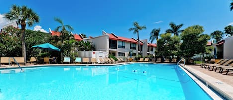 A clear swimming pool with lounge chairs and umbrellas, surrounded by palm trees and buildings with red roofs.