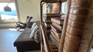 A cozy reading nook with a cushioned chair, a stack of books, and a large window with a view of greenery. - Holliday Villa - Casa do Sobreiro (Melres, Gondomar)