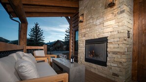 Un área de estar al aire libre acogedora, con chimenea de piedra, muebles de madera y vistas a una montaña y árboles.