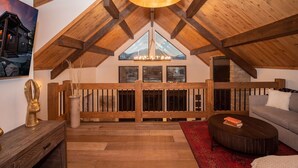 A spacious loft with a high ceiling, wooden beams, a large window with a mountain view, a cozy seating area with a sofa and a coffee table, and a decorative head sculpture on a wooden console. - Rocky Mountain Escape - Steps to Lake Estes, Jacuzzi, Close to Downtown Estes (Estes Park)