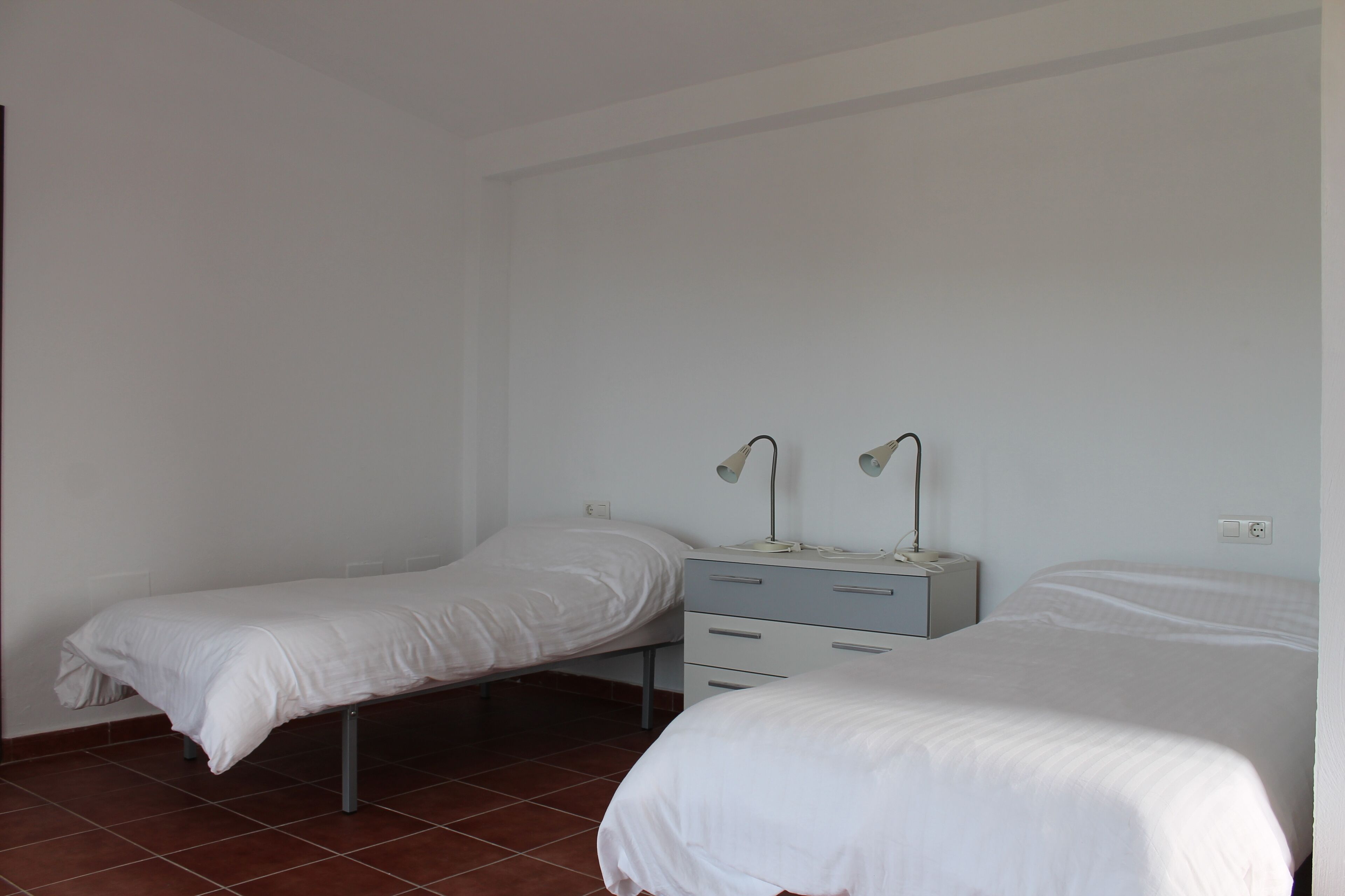 Une chambre simple et bien rangée, avec deux lits simples, un petit bureau avec des tiroirs et une lampe.