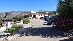 A building with a tiled roof, a parking area with cars, and a mountainous backdrop. - Casa Arts (Alcaucin)