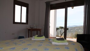 A bedroom with a bed, a small table, and a view of the outdoors. - Casa Arts (Alcaucin)