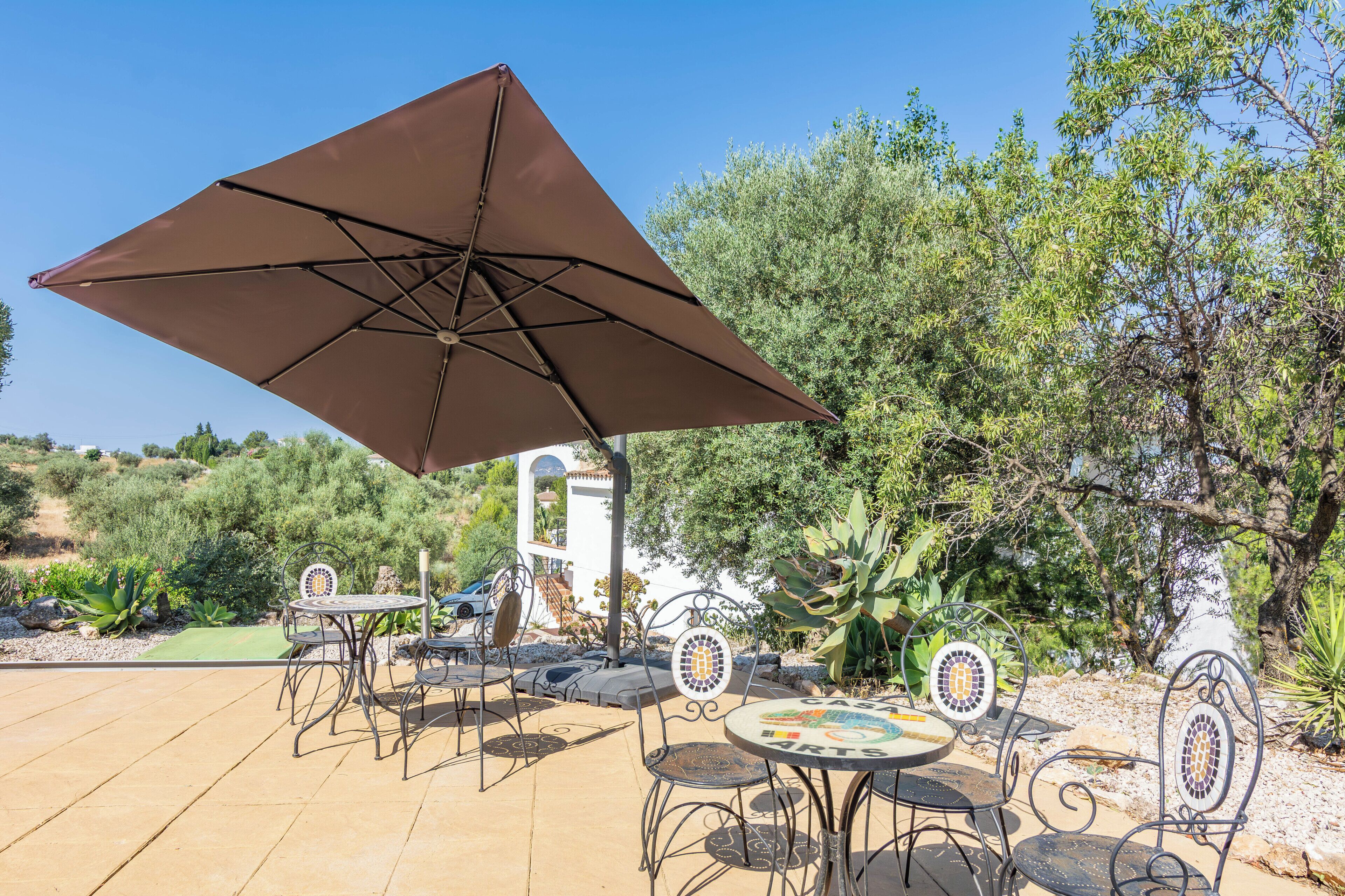 Een terras buiten met tafels en stoelen, een grote parasol en uitzicht op een tuin met bomen en een wit gebouw.