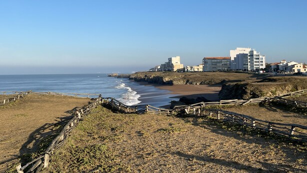 Zona costera con una playa de arena, afloramientos rocosos y una valla de madera.