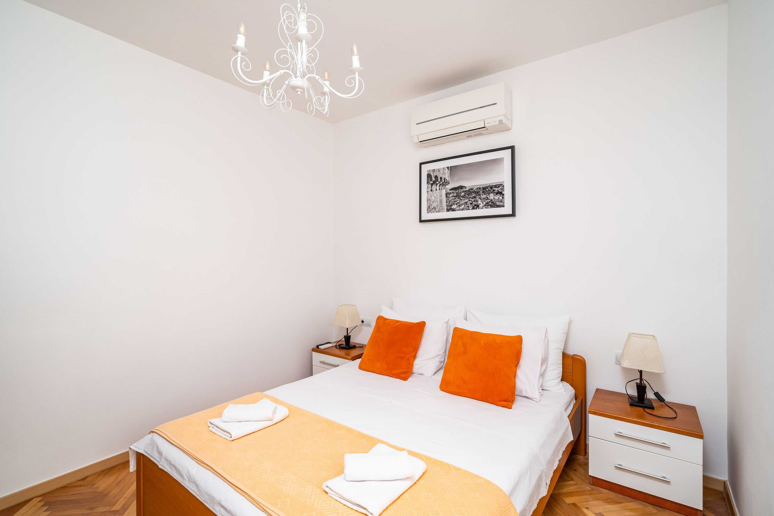 A bedroom with a bed, two nightstands, a chandelier, an air conditioner, and a framed picture on the wall.